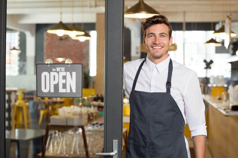 Smiling man with an apron standing next to door of restaurant with open sign