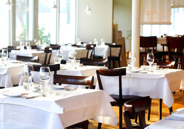 An empty restaurant dining room with many small tables set with white tablecloths, wine glasses, and place settings.