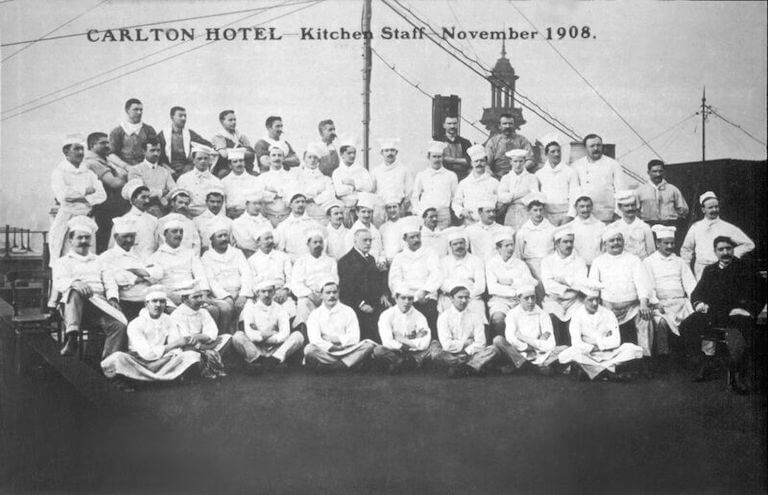 Approximately 60 chefs, nearly all wearing white uniforms, pose for a group portrait; at the center of the group is Escoffier, dressed in black. Text printed on the image states “Carlton Hotel Kitchen Staff, November 1908.”