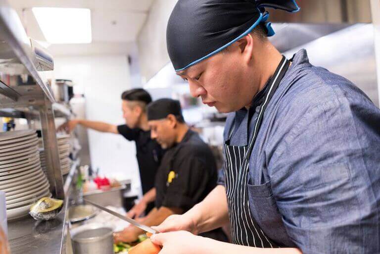 Line cook wearing a striped apron and a black bandana cuts a vegetable next to two other cooks