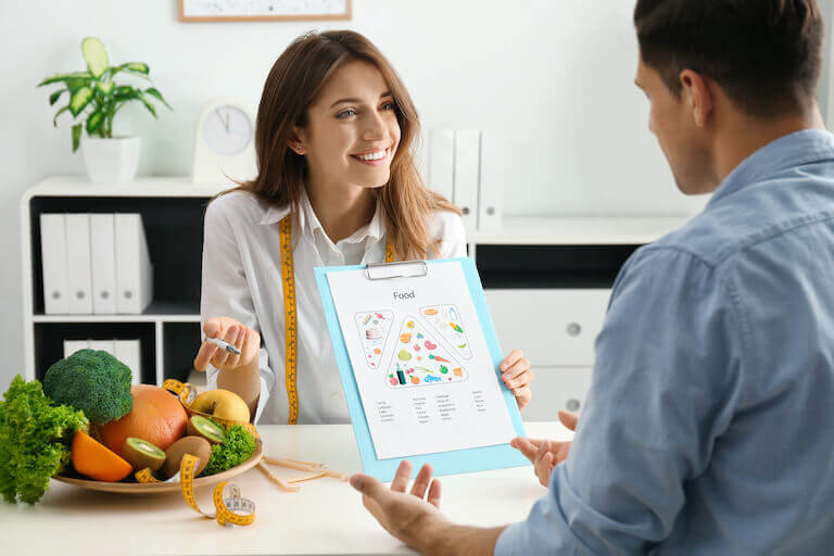 Nutritionist sitting at a desk with a bowl of fruit and vegetables speaking with a patient while showing them a food chart