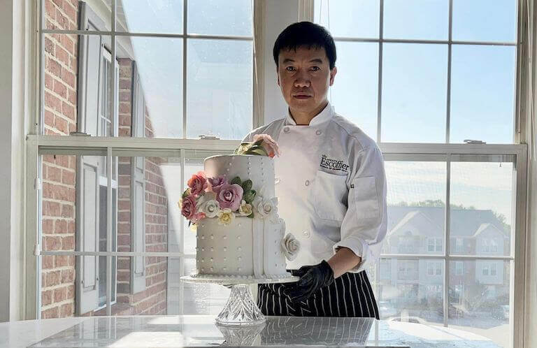 A person in chef's coat stands behind a glass top table with a two-tiered wedding cake on a display stand. The cake features fondant, floral decorations, and piping.