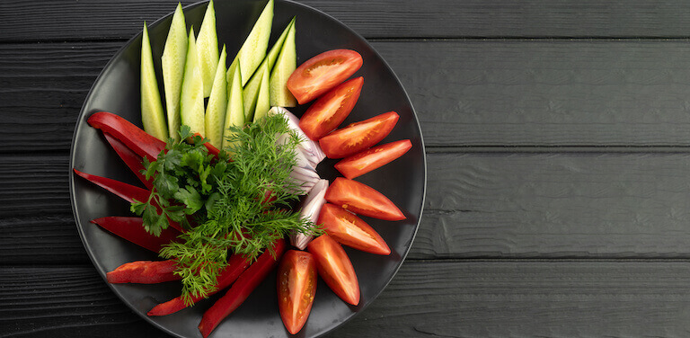 A black bowl of bias cut veggies sits on a countertop, including tomatoes, cucumber, and onions.