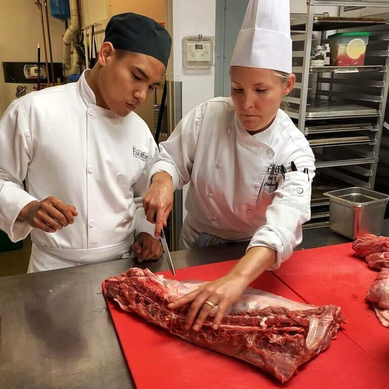 Chef Instructor standing next to a student demonstrating whole-hog butchery