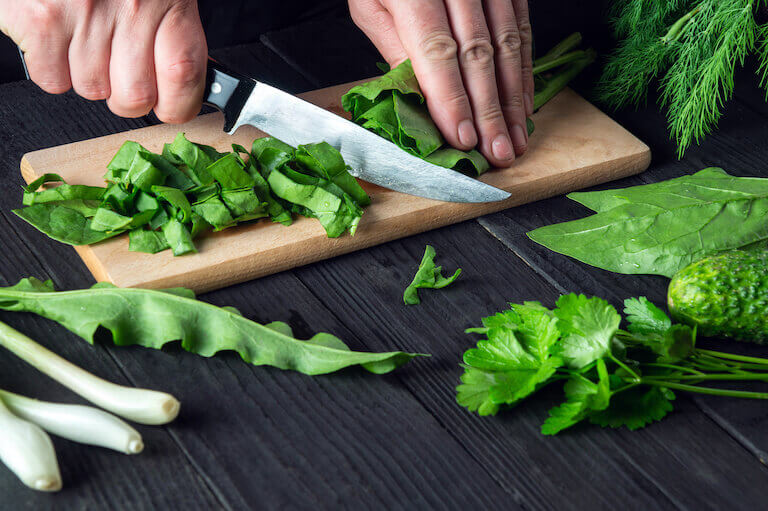 A pair of hands holding a small knife and slicing into bright green herbs rolled up tightly to create ribbons.