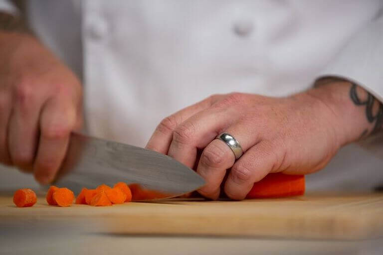 Closeup of a pair of hands cutting a carrot with a chef’s knife.