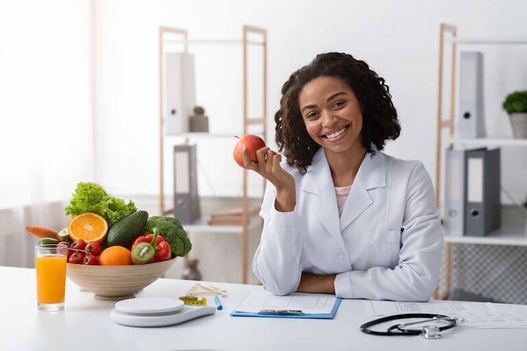 Doctor in uniform sitting at a table holding an apple with a bowl of fruit and vegetables on the table in front of them