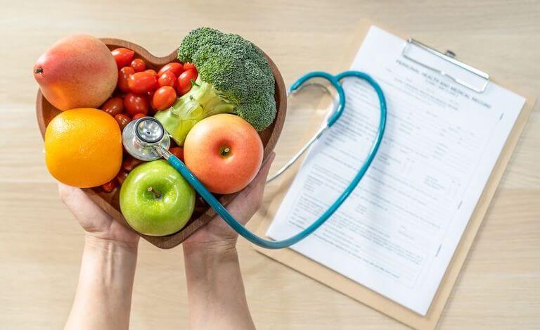 Hands holding a heart shaped bowl filled with a stethoscope, apple, oranges, and other fruits and vegetables