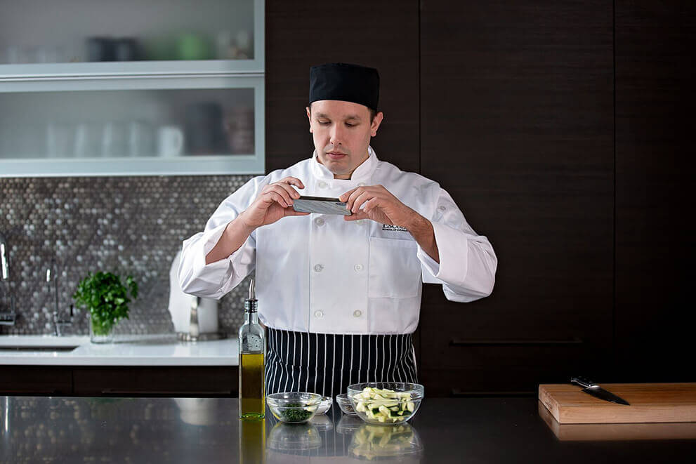 A culinary student in a white chef’s coat and black hat photographs bowls of chopped ingredients on a kitchen counter, documenting mise en place.