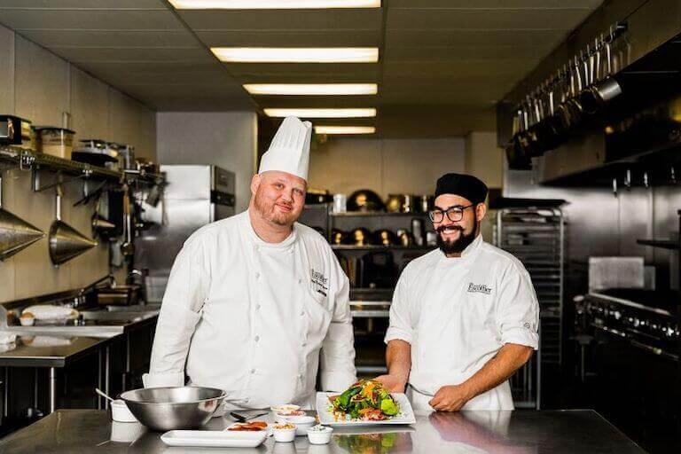 A smiling Escoffier student wearing a white Escoffier uniform displays a dish in a professional kitchen environment with a Chef Instructor by their side.