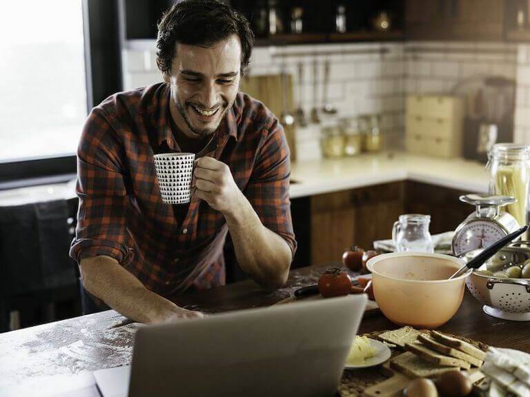 Man holding a mug while using a laptop on the kitchen counter