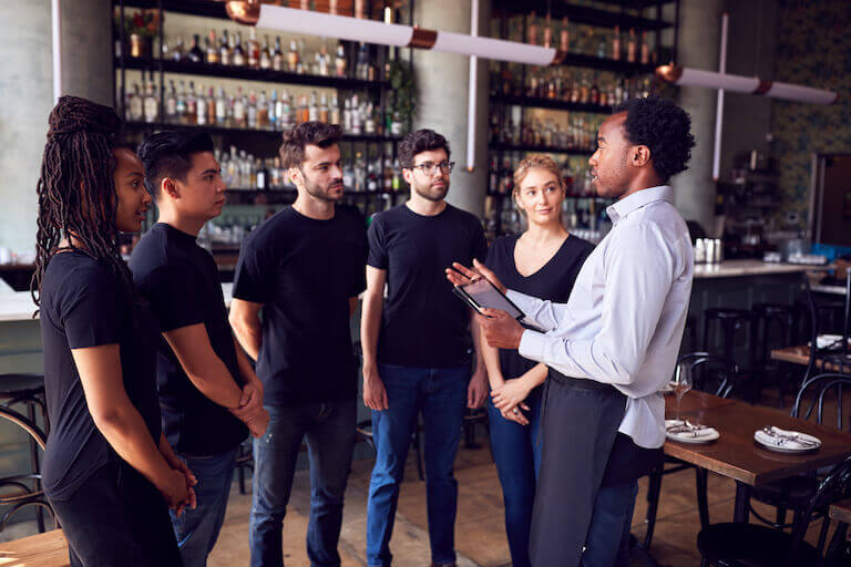 Male manager talking to a team of five staff members, all wearing aprons and standing in front of a restaurant bar