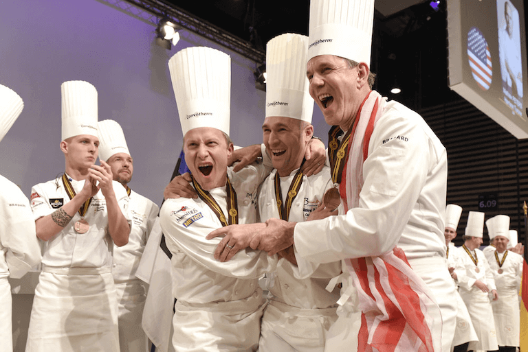 Three chefs in uniform smiling and celebrating on stage with medals