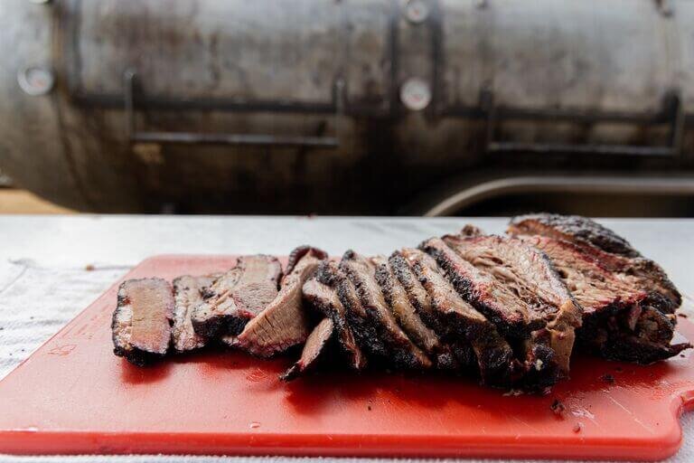 Slices of smoked beef brisket with a dark bark, arranged on a red cutting board with a large metal offset smoker blurred in the background.