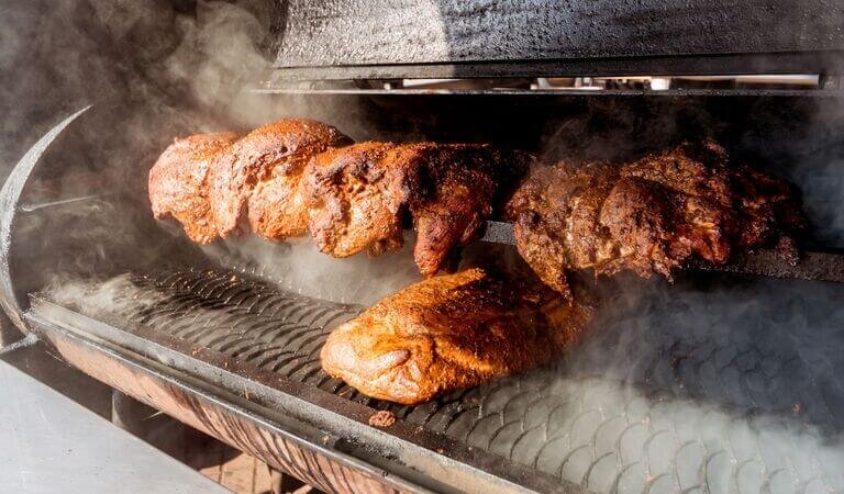 Large, seasoned cuts of meat smoking inside an open black commercial barbecue smoker, with thick smoke rising around the food.