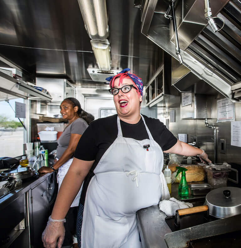 Chef with pink hair wearing a white apron smiling as they work in a food truck