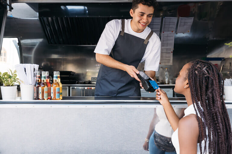A smiling worker stands in the window of a food truck and helps a customer pay for their order with a credit card.