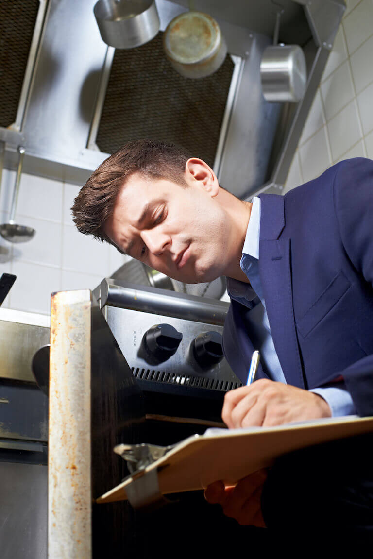 Health inspector writing on a clipboard while inspecting an oven in a commercial kitchen