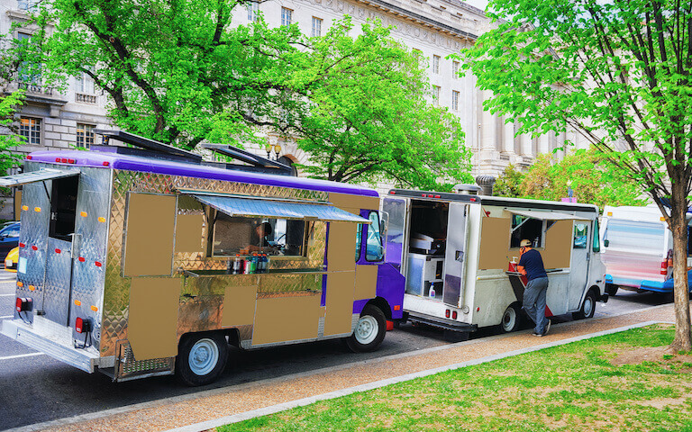 Two food trucks parked on a street under trees