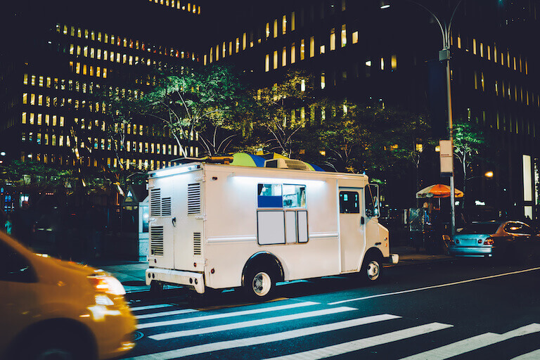 A white food truck parked by the curb in an urban setting at night, with a small park and a tall building with many lights on in the background.