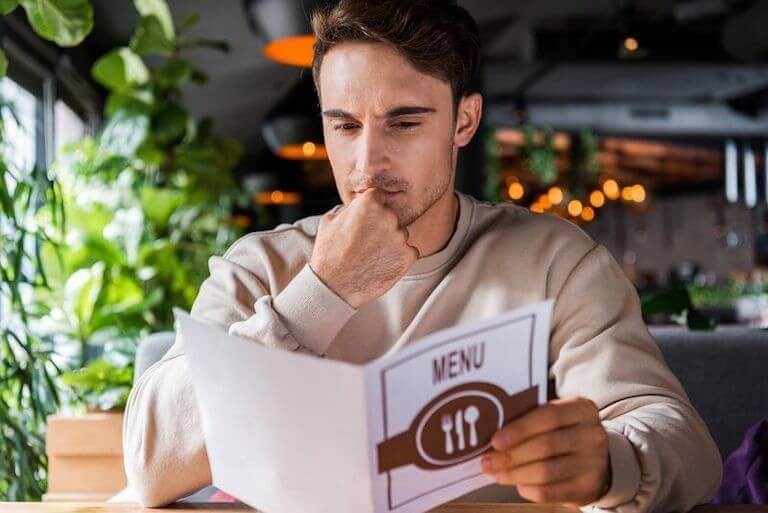 A customer sits in a restaurant booth near a bright window decorated with lots of fresh plants and considers the contents of a menu in their hands.