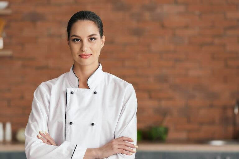 Chef wearing a white uniform with her arms crossed in a kitchen