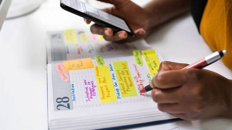 Person writing goals in a planner, while holding a cell phone.