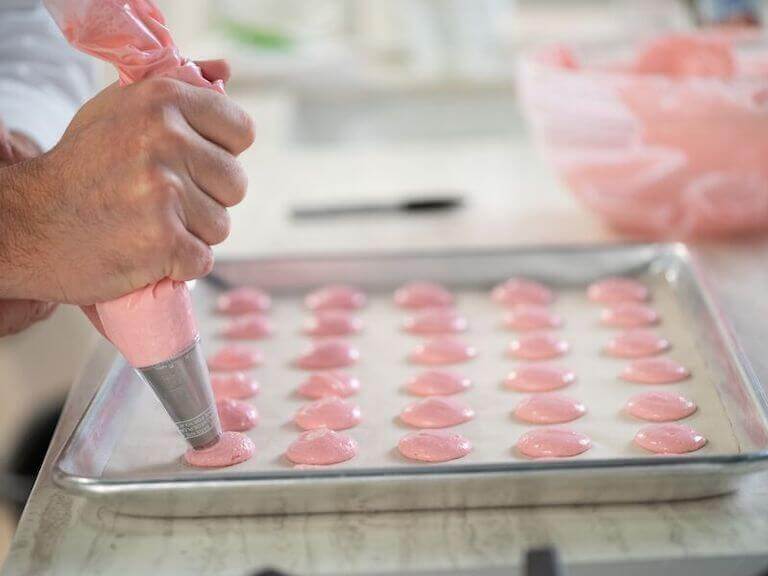 Close up of a bakers hand piping pink macarons on a baking sheet