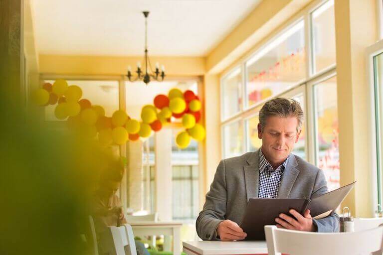 A well-dressed man reads a large brown menu in a bright, cheerful restaurant.