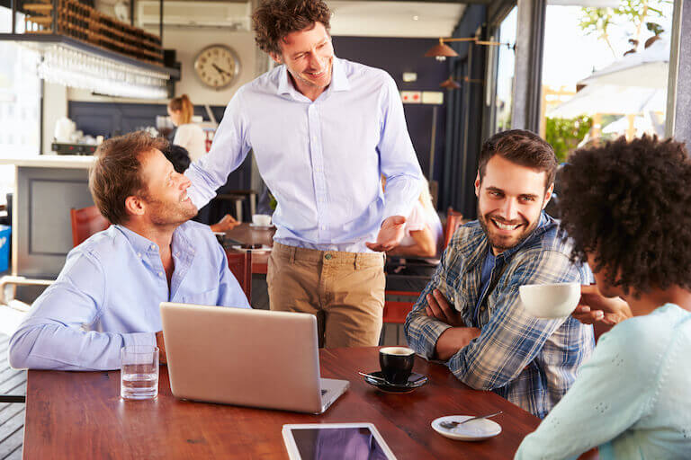 A restaurant manager chats with three smiling patrons at their table in a casual restaurant environment.