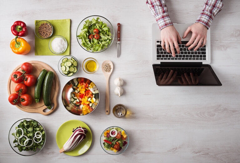 Top view of hands typing on a laptop next to cut vegetables on the table