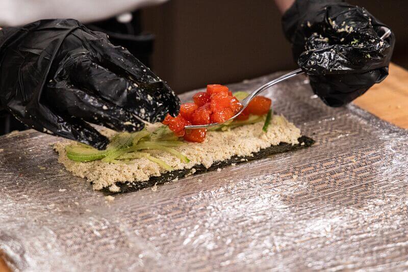 Close-up of a cook wearing black gloves assembling a sushi roll, spreading seasoned rice over nori and adding sliced cucumber, avocado, and diced salmon on a plastic-wrapped bamboo mat.