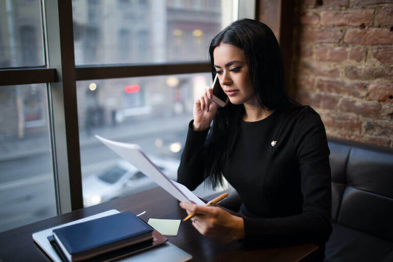 A female business owner sits near a second floor window talking on the phone and holding a paper.