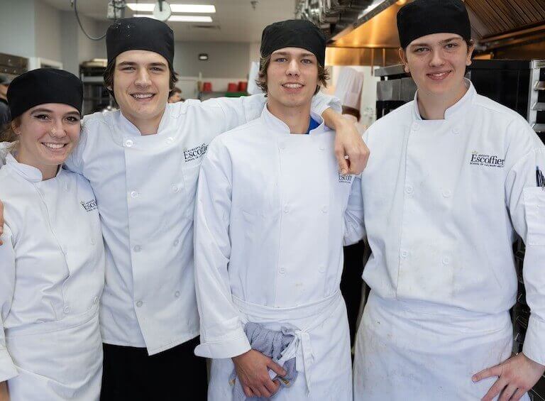 Four Escoffier students in white uniforms posing for a photo together in a kitchen