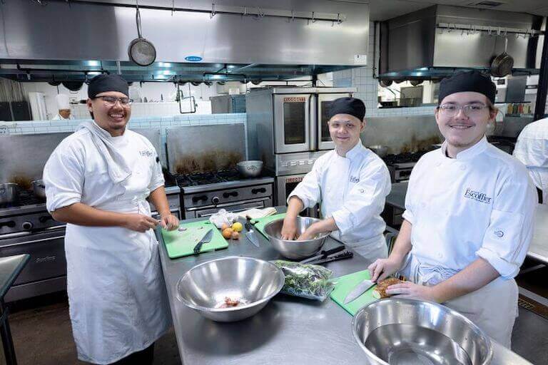 Three Escoffier students making salads in large metal bowls in a kitchen