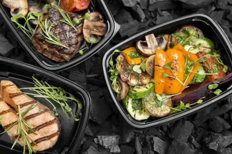 Three black meal prep containers filled with grilled salmon, steak, and colorful roasted vegetables, garnished with fresh herbs and microgreens, arranged on a bed of charcoal.