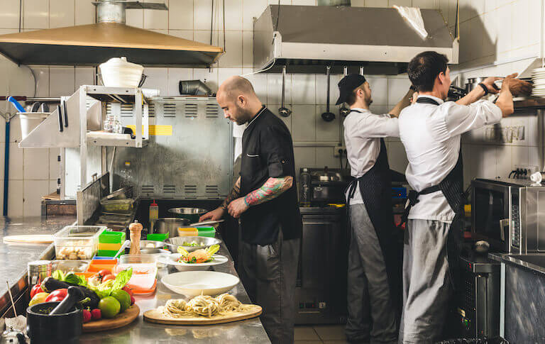 A cook in a black uniform plates dishes in a restaurant kitchen while, nearby, two chefs in white uniforms and black aprons stack dishes on a shelf.