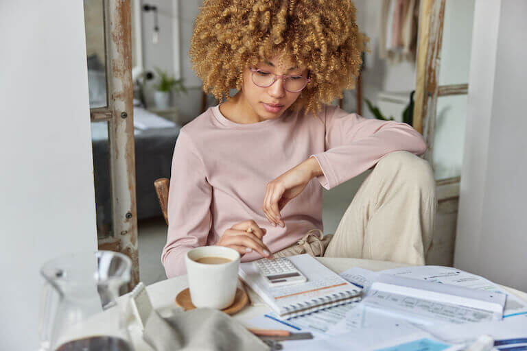 Person with curly hair sitting at a table cluttered with papers while using a calculator