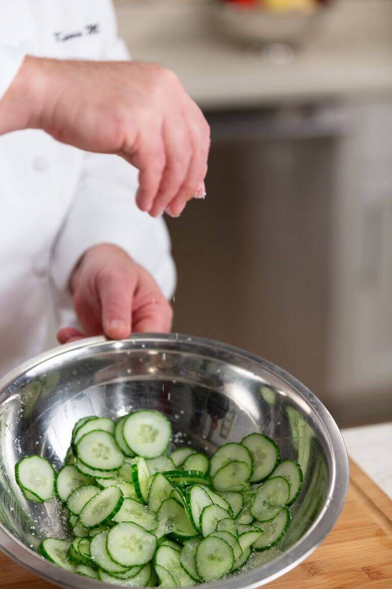 Chef salting cucumbers in a metal bowl