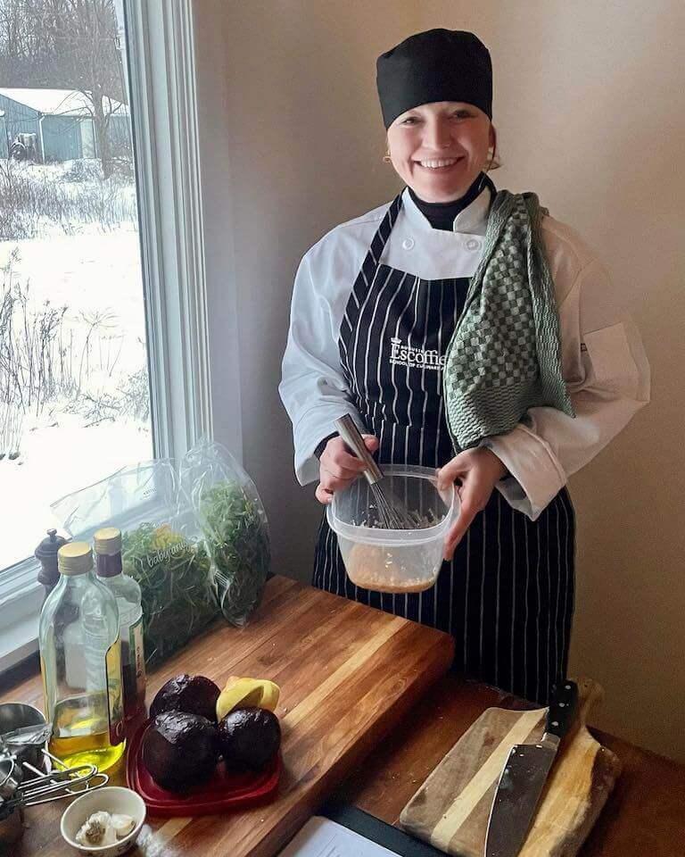 A student in a white chef’s uniform with black-and-white striped apron smiles into the camera and holds a whisk and a plastic container. They are standing next to a window inside a house with an array of ingredients on a small wooden table in front of them.