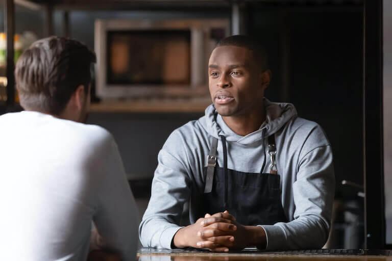 A chef in a black apron talking with a man at a table.