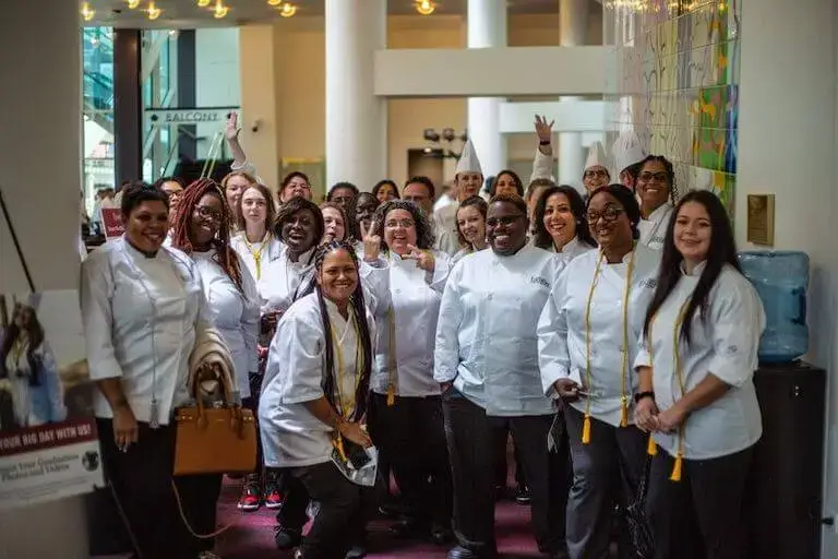 Group of diverse Escoffier graduates in chef whites and hats celebrating at graduation ceremony
