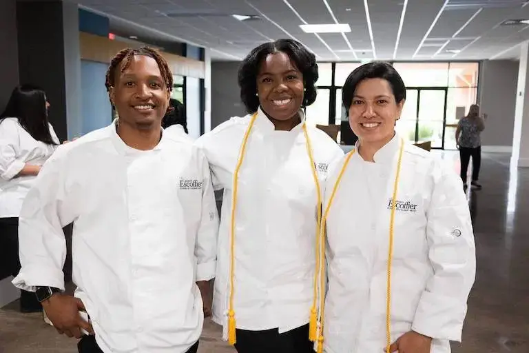 Three diverse Escoffier graduates in chef whites and graduation stoles smiling together at graduation ceremony