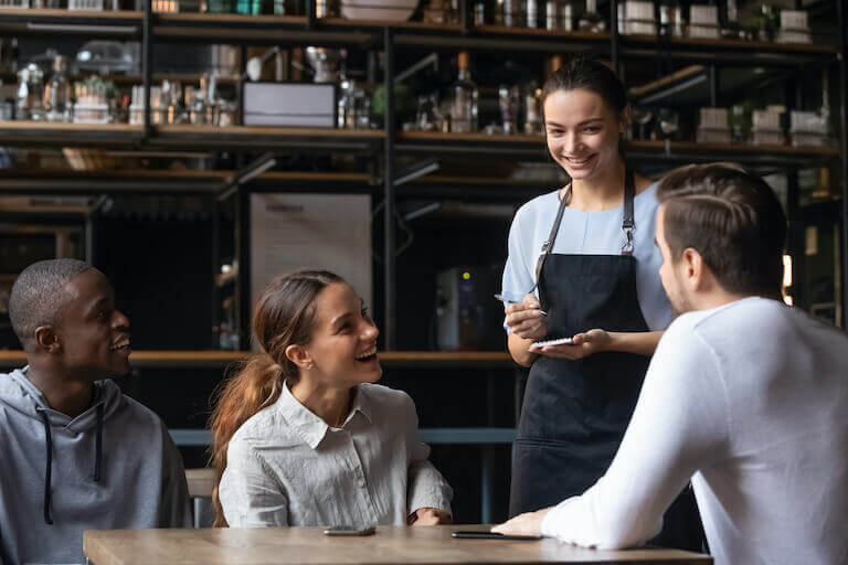 A smiling server chats with three happy patrons in a modern restaurant dining room.