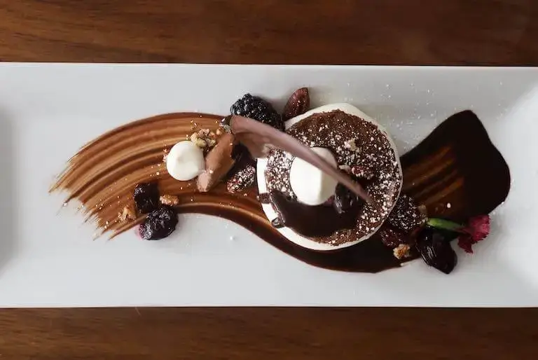 An overhead view of an elegantly plated chocolate dessert on a white rectangular plate, featuring a small round cake, fresh berries, pecans, cream dollops, and a decorative swirl of chocolate sauce.