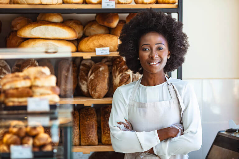 Baker wearing a white apron standing in front of fresh bread with arms crossed