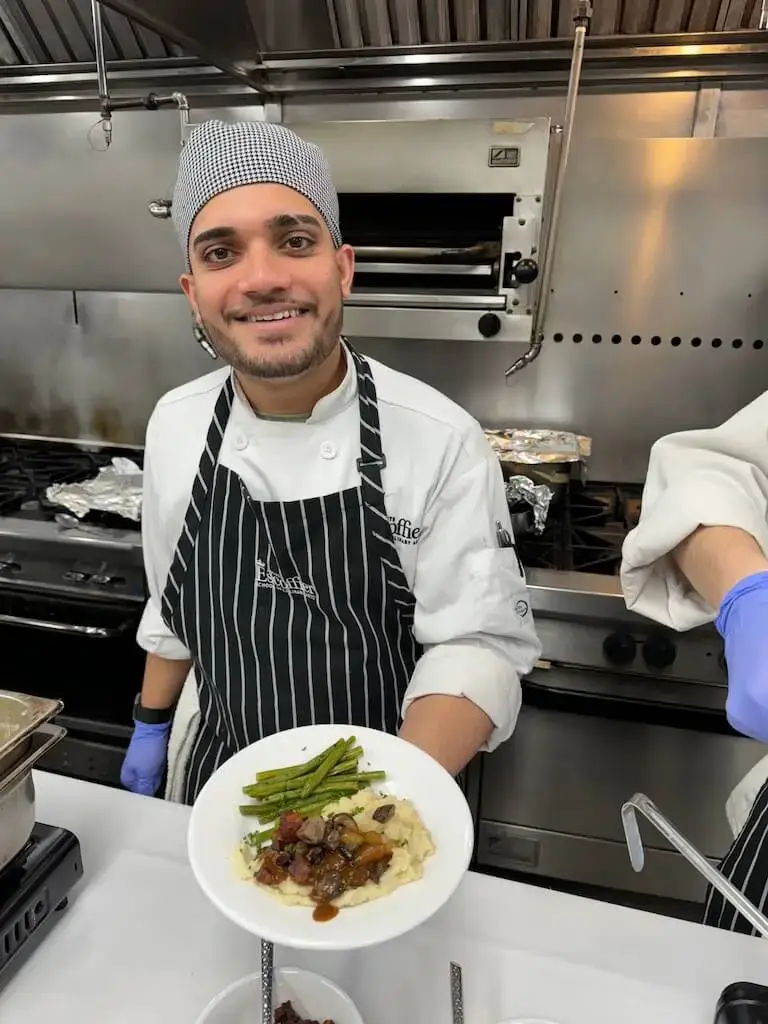 Escoffier student in white chef coat and striped apron holding a plated dish with protein, vegetables, and sauce in a commercial kitchen