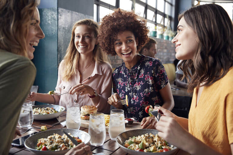A group of young women smiling and laughing over their entrees in a casual restaurant setting.