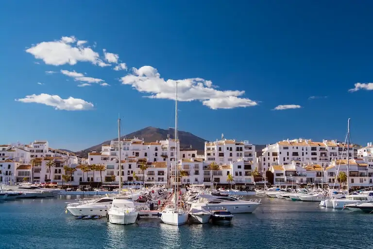 Yachts docked in a marina with white buildings and mountains in the background