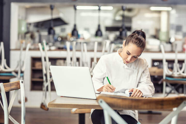 A chef sits in an empty restaurant, with chairs turned over on the tables, writing in a notebook with their laptop open.